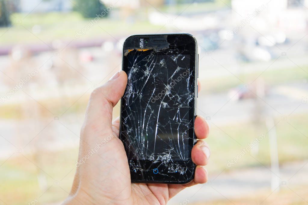 Broken glass screen smartphone in hand of upset man. The guy is holding a black smartphone with a broken display. Broken screen of modern gadget. large crack in the form of a web on the smartphone