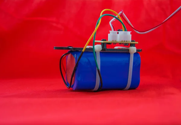 Part of an electronic device. Blue battery pack with printed circuit Board fastened by zip tie, connectors and colored wires on red background.