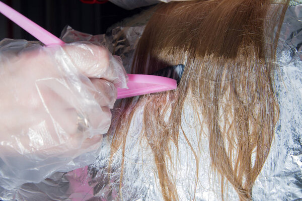 Hairdresser makeup artist hand in a transparent disposable glove applies hair dye with a pink brush on the hair of a blonde. Hair coloring process, highlighting