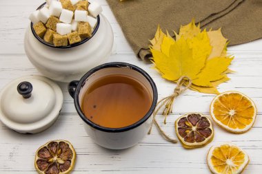 Alüminyum fincanda çay, alüminyum şeker kasesinde şeker, kahverengi eşarp, akçaağaç yaprakları, kuru portakallar ve açık ahşap arka planda kurutulmuş limon.