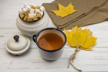 Alüminyum fincanda çay ve alüminyum şeker kasesinde şeker küpleri, kahverengi eşarp, açık ahşap arka planda akçaağaç yaprakları.