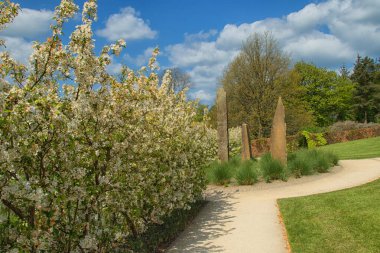 Garden path lined with White Apple blossom leading to tall thin carved stone sculptures, RHS Garden, Harlow Carr, Harrogate, North Yorkshire, England, UK. clipart