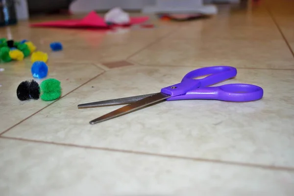 Multi colored cotton pompoms, glue, glue sticks, construction paper, and scissors covering a linoleum floor during arts and crafts time