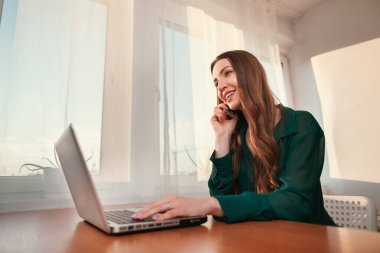 Şirket iletişimi. Modern ofiste iş kadını cep telefonuyla konuşuyor. Genç, mutlu, iş kadını ofiste dizüstü bilgisayar kullanarak oturuyor.. 