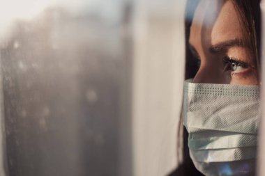 Coronavirus. Covid-19 'un hasta kadını pencereden bakıyor ve maske takıyor. Hasta karantina sırasında enfeksiyonu önlemek için izole edilmiş. Kadın hava kirliliği yüzünden maske takıyor..