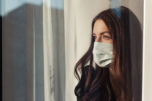 Coronavirus. Covid-19 'un hasta kadını pencereden bakıyor ve maske takıyor. Hasta karantina sırasında enfeksiyonu önlemek için izole edilmiş. Kadın hava kirliliği yüzünden maske takıyor..