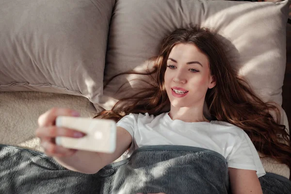 Beautiful girl making selfie in the bed. Top view of young woman in white shirt making selfie using a smartphone and smiling.