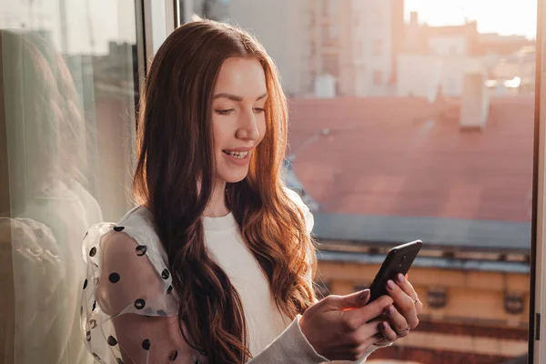 Gülümseyen beyaz kadın pencere eşiğinde oturuyor ve evdeki akıllı telefona mesaj yazıyor. İnternet 'e bağlanmak ve sosyal medyayı mutlu etmek için akıllı telefon kullanan genç bir kadının yan görünümü.