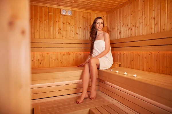 Genç ve güzel bir kadın saunada dinleniyor. Vücut bakımı kavramı. Genç bir kadın saunada ahşap bankta oturuyor..