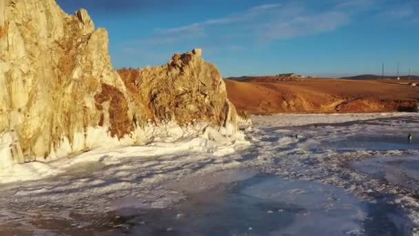 Zamarznięte jezioro Baikal, skała szamańska Cape Burhan na wyspie Olkhon. Turyści nad jeziorem Baikal, spacerujący po lodzie jeziora.. Słynna krajobrazowa Rosja. Niebieski przezroczysty lód z głębokimi pęknięciami. — Wideo stockowe