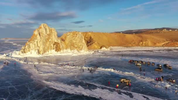 Zamarznięte jezioro Baikal, skała szamańska Cape Burhan na wyspie Olkhon. Turyści nad jeziorem Baikal, spacerujący po lodzie jeziora.. Słynna krajobrazowa Rosja. Niebieski przezroczysty lód z głębokimi pęknięciami. — Wideo stockowe