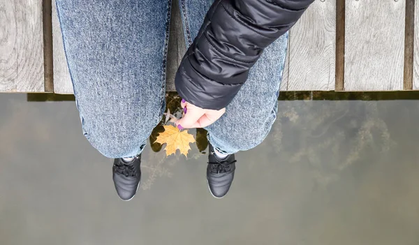 Mooie benen van een meisje in jeans zitten op een houten brug bij het meer, met een geel esdoornblad in hun handen. Herfst warme en zonnige dag. Zicht van bovenaf. Najaarsconcept — Stockfoto