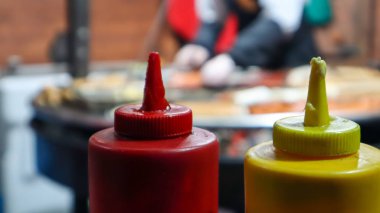 Ketçap ve mayonez plastik borularda ön plandaki ızgaranın yanında seçici bir odak ile. Barbekü, sokak yemeği festivali. İki kutu et sosu. Kırmızı ve sarı konteyner.