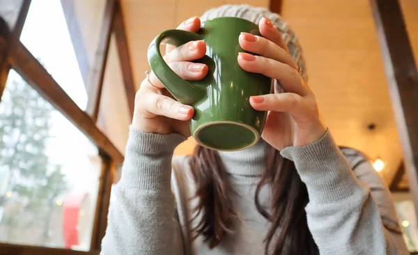 Acercamiento de las manos femeninas con una taza de bebida. Una chica en un suéter gris sostiene una taza de café o cacao en un café en la mesa y mira por la ventana. Bebida caliente. Una foto acogedora con un fondo borroso —  Fotos de Stock