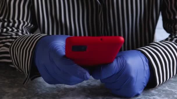 女人的手戴着蓝色防护手套的特写 用红色的手提箱在智能手机上输入短信 坐在桌子旁边 以免感染考拉病毒 保护措施 — 图库视频影像