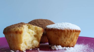 Çikolata ve krema serpiştirilmiş üç kremalı kek, biri ısırılmış, pembe bir tabakta, mavi arka planda, yakın planda. Tatlı, küçük bir kek. Havadar desenli beyaz kurabiyeler.