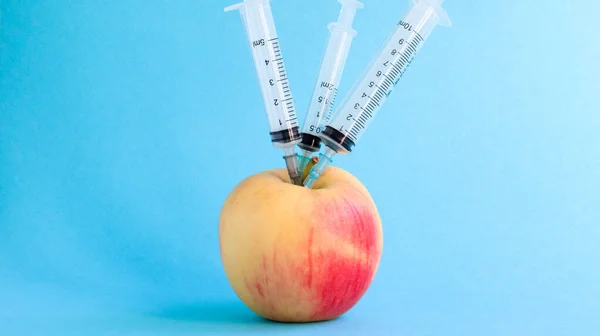 Three different medical syringes sticking deep into an apple on a blue background. The fruit is strewn with syringes. A ripe red apple pierced from all sides by syringes. Healthcare or cosmetology