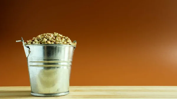Small metal bucket with raw buckwheat on a board on a brown background. Organic vegetarian food. The concept of a healthy and balanced diet. Dietary nutrition. Copy space.