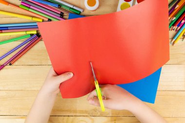 Çocuk renkli kağıtları makas ve tahta arka plan üzerine çizim yapmak için bir sürü kalem ve boya ile kesiyor.