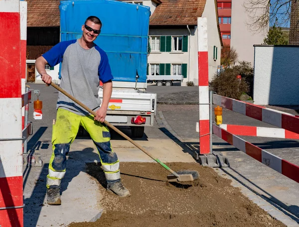 Trabajador de la construcción riendo con escoba de pie en la arena delante del vehículo y el remolque mirando a la cámara . — Foto de Stock