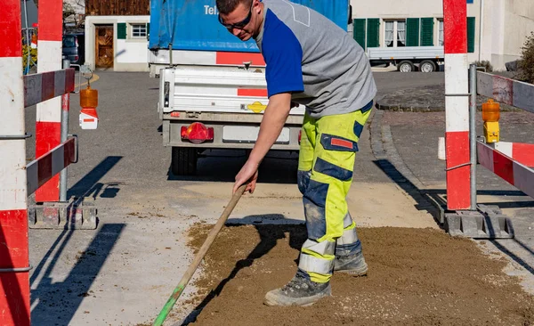 Trabajador de la construcción con gafas de sol y escoba limpiando arena en el sitio de construcción delante de un vehículo de construcción y remolque . — Foto de Stock