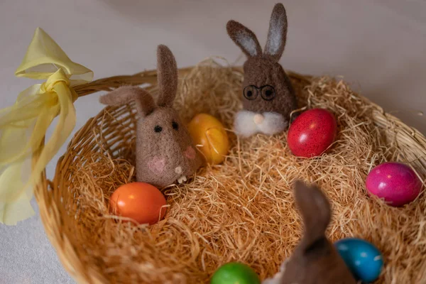 Colored easter eggs and caps of brown felt bunnies in nest of straw in straw basket. Felt bunnies as warm holder for easter eggs.