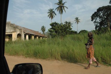 Walking African woman on street of sand with high green meadow, palms and an African church in background. wearing brown colored clothes with African pattern. View throughout the window of a car. 11 February 2018 Pointe Noire, Congo. clipart