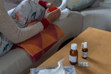 Kadın ilaçla çay içiyor ve oturma odasında kanepede oturuyor. Hasta hissetmenin konsepti.
