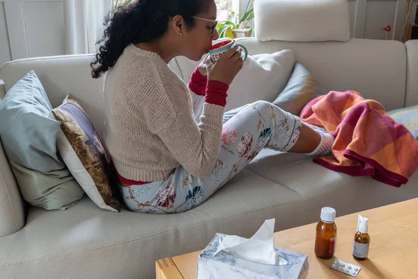 Woman having a cold, sitting on white couch in the living room while drinking tea. Legs in white warm socks, medicine and tissue paper on table. Concept.