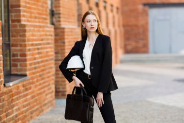 Kızıl saçlı bir mimar kız elinde koruyucu bir miğferle tuğla bir binanın arka planında duruyor ve yüzünü çeviriyor.