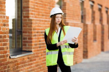 Koruyucu miğferli, mutsuz, kızıl saçlı bir mimar kız tuğla bir binanın önünde elinde çalışma belgeleriyle duruyor ve tatmin olmuyormuş.