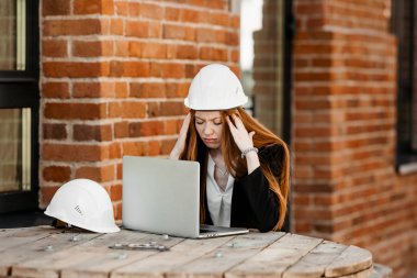 Kızıl saçlı bir kız koruyucu kasklı bir mühendis ofiste dizüstü bilgisayarın başında oturur ve yorgunluktan ve baş ağrısından ellerini başının üstüne koyar.