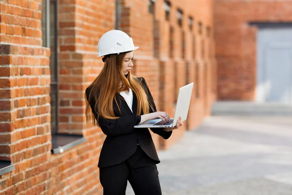 一位戴着防护头盔的红头发建筑师女孩站在一座砖楼前 手里拿着笔记本电脑 不悦地看着显示屏 — 图库照片