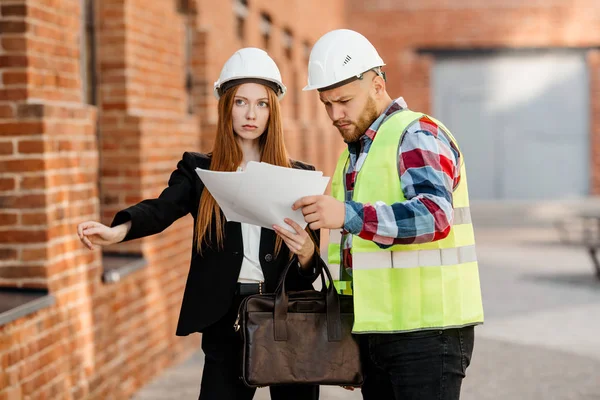 一个红头发的年轻女孩 戴着防护头盔的工程师 与一名施工工头讨论工作文件 — 图库照片