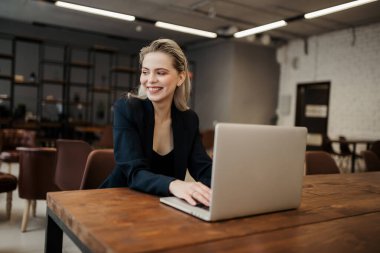 Klasik takım elbiseli ekonomist bir kız iş yerindeki bir bilgisayarıyla masaya oturur ve gülümseyerek pencereden dışarı bakar.