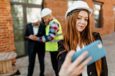 Koruyucu miğferli kızıl saçlı bir mimar kız elinde bir telefonla meslektaşlarının arka planına bakarak iyi koordine edilmiş bir takım çalışmasını fotoğraflıyor.