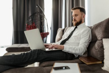 Gömlek ve kravat takmış sakallı bir erkek serbest olarak kanepeye yayılmış, oturma odasında oturuyor ve bir dizüstü bilgisayar üzerinde çalışıyor.
