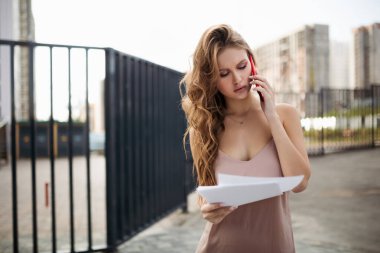 İş kadını çalışan belgeleri tutuyor ve yüksek binaların yanında duruyor. Telefonda önemli bir iş görüşmesi yapıyor.