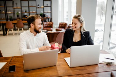 Sakallı bir adam ofis masasında oturuyor ve iş yerindeki güzel meslektaşına bir hediye veriyor.