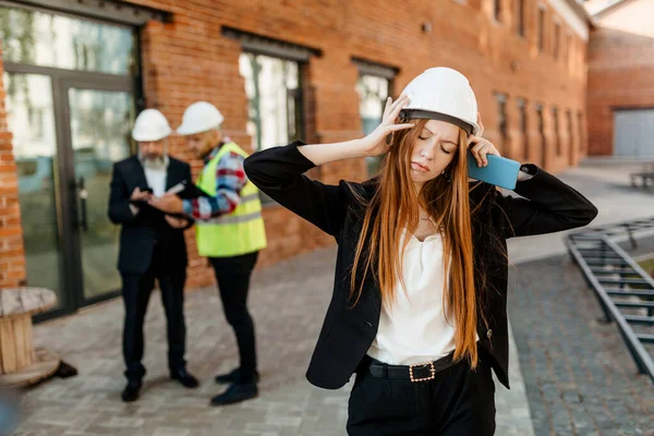 一个戴着防护头盔的红头发建筑师女孩站在一群工人面前 由于头疼和疲倦而把头抬起来 — 图库照片