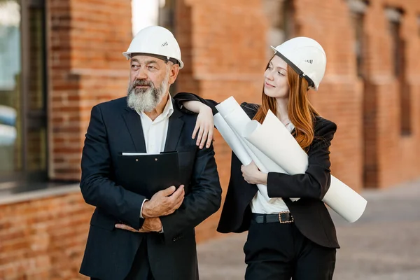 一个戴着白色防护帽的红头发女工程师和一个穿着西装的白头发老年建筑师代表了团队合作 — 图库照片