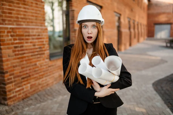 A red-haired engineer girl in a White protective helmet stands on the street in front of the office, holding sketches and drawings. And her face shows surprise