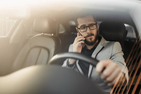 Бородатый бизнесмен в очках сидит за рулем представительской машины и разговаривает по телефону. Фото через лобовое стекло
