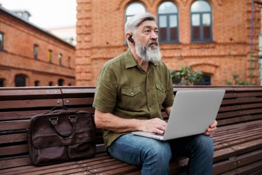 Beyaz saçlı bir iş adamı öğle yemeği sırasında parkta oturur ve bilgisayarındaki son haberleri inceler.