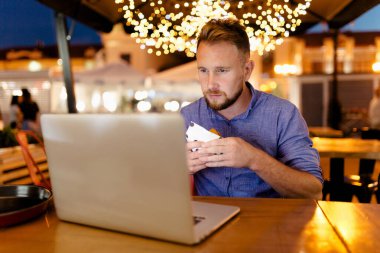 Sakallı bir erkek serbest yazar akşamları bir kafede oturup hamburger yer ve dizüstü bilgisayarla çalışır. Arkaplanda güzel bir ampul topağı var.