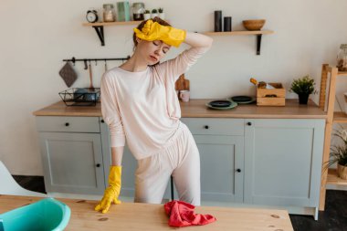 Sarı lastik eldivenli kızıl saçlı kız, mutfakta duruyor ve evi temizlemekten dolayı yorgunluk çekiyor.