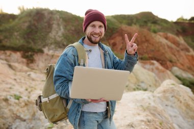 Sakallı bir hippi elinde dizüstü bilgisayarla duruyor ve barış jesti yapıyor.