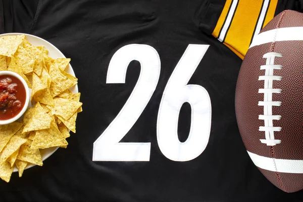 An American football with organic nacho chips and mild salsa on a white black football jersey with the 26 number on horizontal view — 스톡 사진