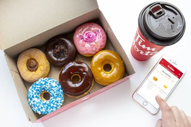 Calgary, AB Canada. Jan 27 2020. A person using an iPhone using the Tim Hortons app with a coffee and a box of donuts with a Dulce de Leche, Chocolate Truffle, Strawberry, Vanilla Dip, Chocolate.