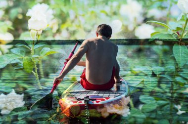 Romantik bir adam Sup tahtasında oturup orman gölünde kürek çekiyor. Beyaz güller ve sörfçü çifte çekim.